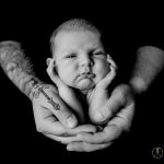 Black and white photo of a newborn boy in his fathers hands. Fathers hands and arms are covered in tattoos. Portrait by Brisson Imagery