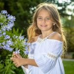 Young girl stands outside holding a blue flower, the sun shining through her golden hair. Portrait by Brisson Imagery