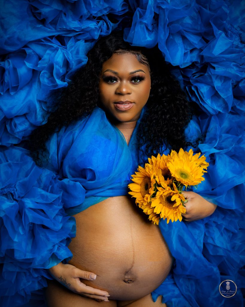 Maternity Portrait by Brisson Imagery. Mother lays with a blue ruffles dress around her head, her belly exposed, she's holding yellow sunflowers.