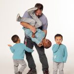 father with 3 young boys, one is dancing around his leg, one he is holding upside down and the other is bashfully smiling at the camera