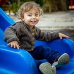 Toddler boy smiling big at the end of a blue slide