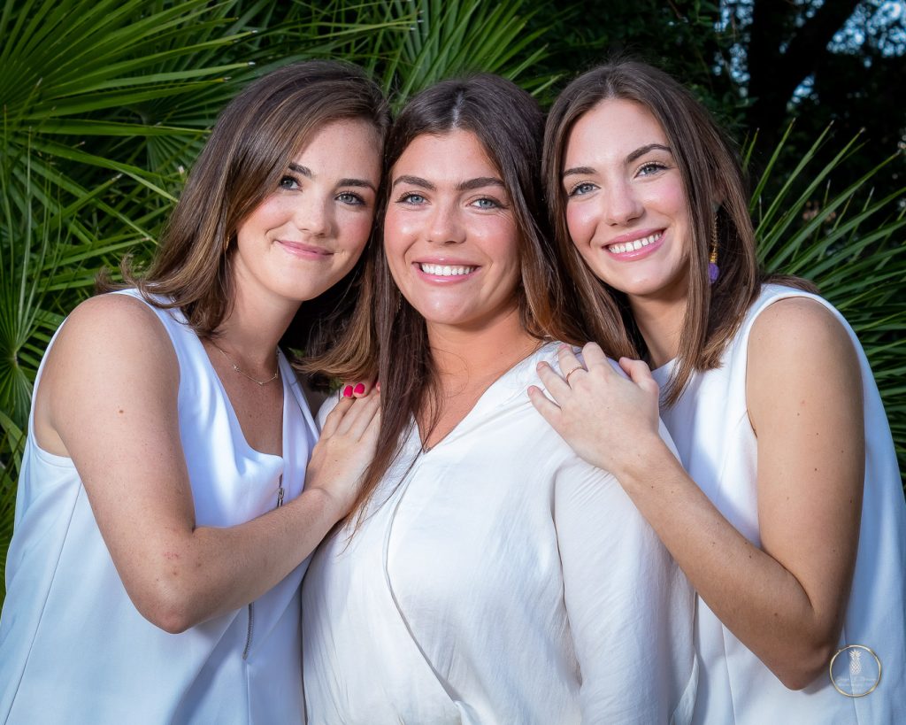 3 adult sisters pose together in a loving embrace. All wearing white. Green palm Frans in the background. Image by Brisson Imagery
