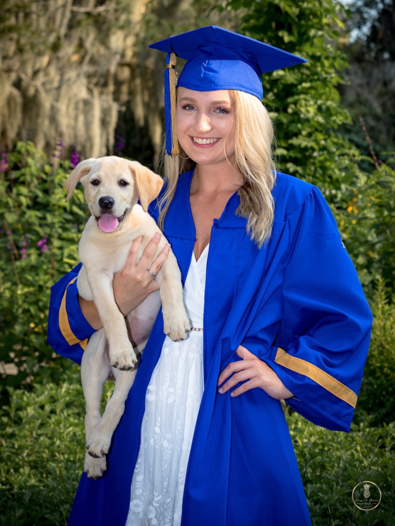 High School Senior poses in cap and gown with a yellow lab puppy in her arms. Portrait by Brisson Imagery