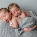 Newborn Twin boys poses on tummies, one on top of the other. Gray backdrop. They still have their hospital bands on. Portrait by Brisson Imagery