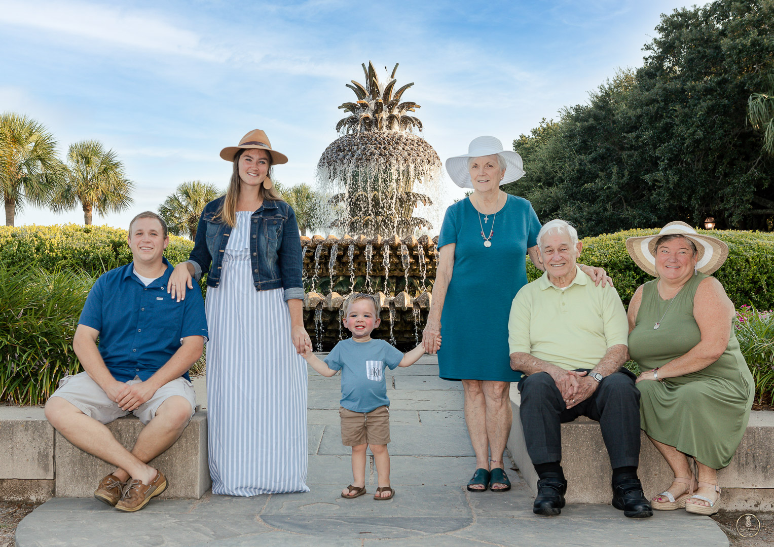 Extended family portrait by Brisson Imagery. Family of 6 lined up in front of Pineapple fountain in Charleston SC. Everyone is color coordinated in blue and green. The woman all have large brim sun hats on. One toddler boy stand in the middle