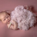 Newborn baby girl on a pick backdrop wearing a fluffy pink tutu and pearls. she is in the reverse taco pose. Portrait by Brisson Imagery
