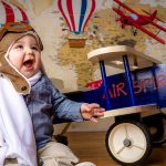 first birthday portrait of a boy wearing a bomber jacket and pilot hat, map as a backdrop, ride on plane next to him