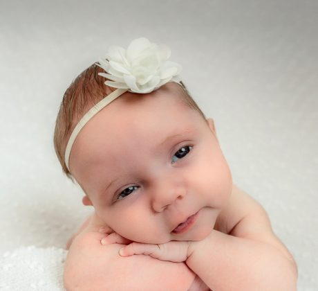 Newborn Baby photo by Brisson Imagery. Baby girl with a white flower headband laying on a white blanket sits with her arms under her chin. Her eyes are open and she is smiling at the camera