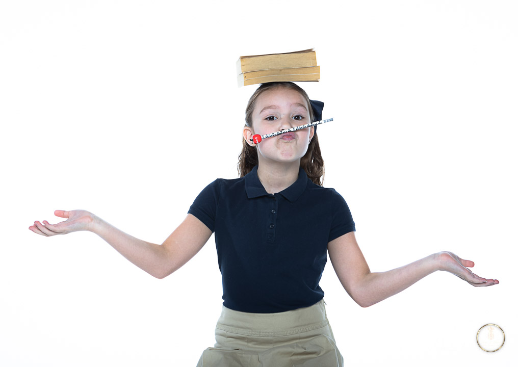 Girl Poses on white backdrop she is wearing a school uniform and is balancing a book on her head and a pencil on her lips
