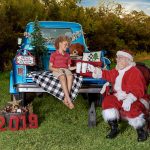 young boy sits on the tail bed of an old blue truck with Santa drinking hot coco