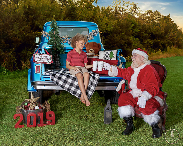 young boy sits on the tail bed of an old blue truck with Santa drinking hot coco