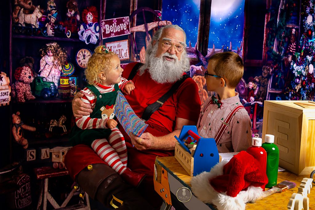 Santa in his workshop with a little girl on his knee and an older boy standing next to him