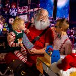 Santa in his workshop with a little girl on his knee and an older boy standing next to him